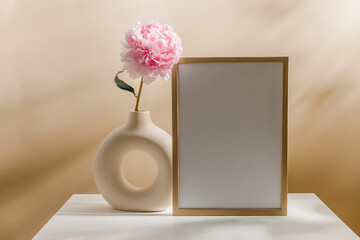 Picture mockup frame with vase and peony flower on beige background with aesthetic sunlight...