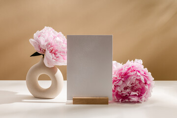 Mockup paper card is standing on wooden paper holder with peony flowers in vase on light brown background with aesthetic shadows. Paper background card for wedding, invitation, branding or design