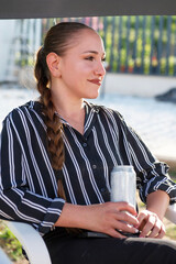 Young woman sitting outdoors, holding a drink in a tin can and looking into the distance with a smile. Relaxing outdoors with your favorite chilled drink in the heat