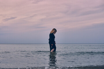 Young woman walks alone in tranquil sea under pastel sky. Her contemplative stance against soothing ocean backdrop evokes introspection and peace