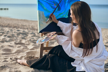 Young woman artist paints by sea, blending colors on palette. Her serene expression reflects...