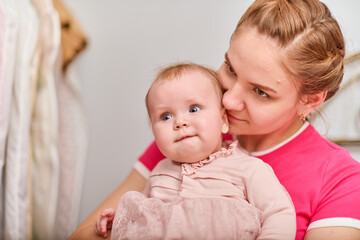 Young mother with light brown hair, holding baby girl. Woman wears pink top, baby in light dress. Warm emotions, indoor environment, bright lighting