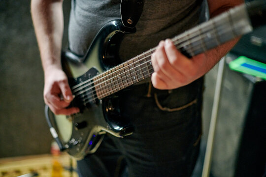 Musician playing an electric bass guitar at music rehearsal, practicing guitar technique in dimly lit setting, close up view guitarist hands