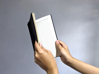 A person's hands holding open a black notebook with lined pages against a plain gray background,...