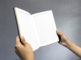 Close-up of two hands holding open a blank lined notebook against a plain grey background,...