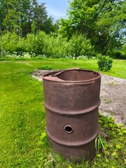 A rusty metal barrel standing in the grass with a garden and green shrubs in the background.