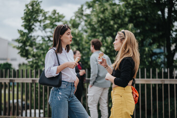 Two young women sharing a casual conversation outdoors in a park-like setting, enjoying ice creams...