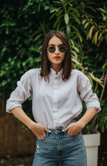 Young confident woman with sunglasses poses outdoors in a stylish white shirt and denim jeans,...
