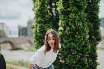 A vibrant young woman wearing sunglasses and casual clothes poses outdoors next to greenery. The...