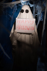 Funny ghost in dark corridor with cobweb showing board with happy halloween inscription positive...