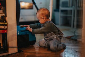 A young child interacts with a vacuum cleaner, kneeling on a wooden floor, featuring a cozy and...