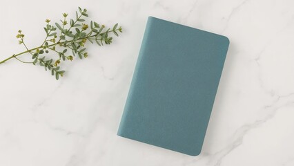 A teal notebook rests on a marble surface, accompanied by a sprig of greenery.