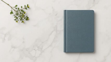 A closed blue notebook rests on a marble surface, accompanied by a small green plant branch.