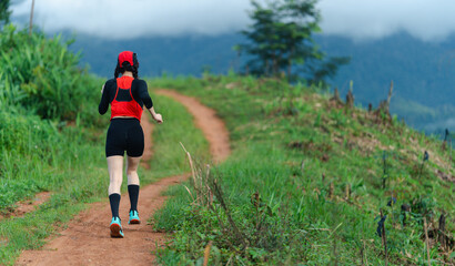 A person jogs on a dirt trail surrounded by green vegetation and distant hills under a cloudy sky.