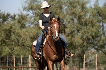 Western lifestyle with cowgirl riding sorrel mare horse in outdoor arena during summer season on ranch.