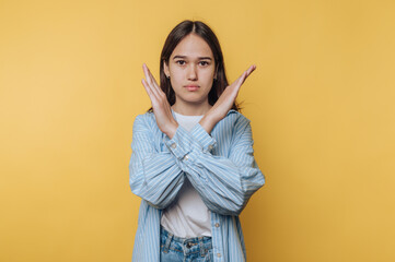 Young woman in casual clothes making an X with her arms against a yellow background.