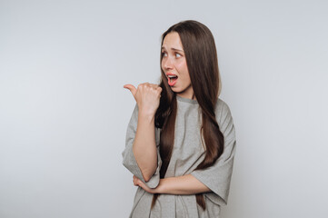 A surprised young woman with long brown hair pointing to the side, wearing a gray shirt, against a...