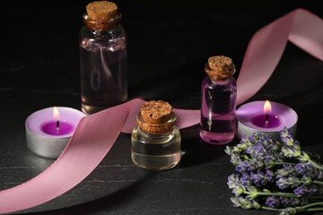 Bottles of lavender essential oil, flowers, burning candles and ribbon on black table, closeup