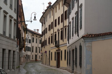 Charming narrow cobblestone street lined with traditional European buildings featuring various window shutters and architectural details, under an overcast sky, evoking a quiet historic town atmospher