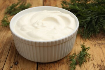 Tasty sour cream in bowl and dill on wooden table, closeup