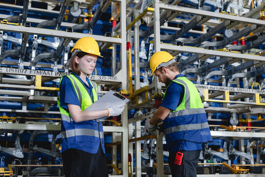 Two industrial workers wearing safety vests and helmets collaborate while inspecting vehicle parts in large auto parts warehouse.
