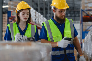 Two warehouse workers wearing safety vests and helmets scanning product barcodes in storage...