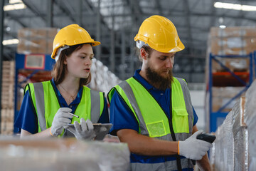 Two warehouse workers wearing safety vests and helmets scanning product barcodes in storage...