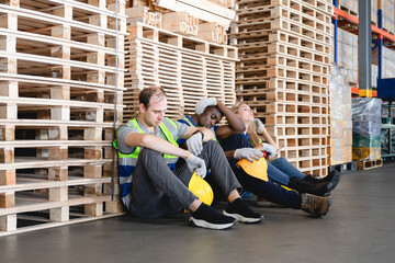 Group of industrial workers in reflective vests taking break near pallet stacks, expressing fatigue...