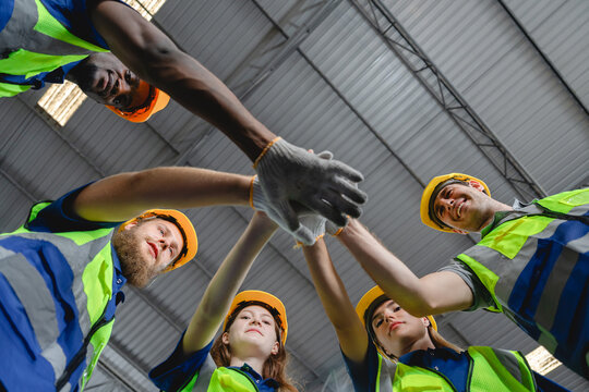 Low-angle shot of multicultural construction workers in safety vests and helmets joining hands in a circle, symbolizing strength, unity, and collaboration in industrial teamwork.
