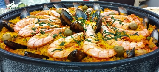 A large bowl of food with shrimp and olives on top. The bowl is filled with yellow rice and the...