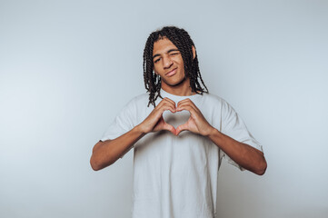 A young man with braided hair making a heart shape with his hands, smiling playfully against a...