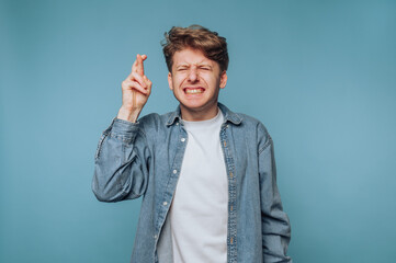 A young man with curly hair crosses his fingers, eyes closed, expressing hope or anticipation...
