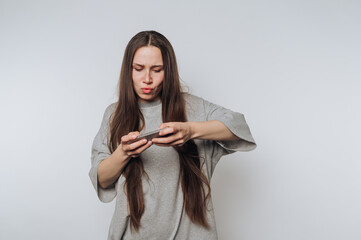 A young woman with long hair, wearing a gray t-shirt, looks intently at her smartphone against a...