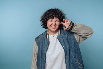 A person with curly hair wearing a denim jacket and white shirt makes a playful gesture against a...