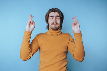 A young man with long hair and a beard, wearing a mustard turtleneck sweater, stands against a blue...