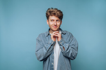 A young man in a denim shirt stands against a blue background, looking to the side with a playful...