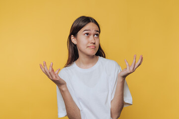 A young woman in a white t-shirt shrugging with a puzzled expression against a yellow background.