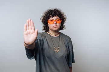 A person with curly hair wearing orange sunglasses and a gray oversized t-shirt, holding up a hand...