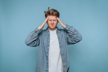 A young man in a denim shirt and white t-shirt stands against a blue background, holding his head...