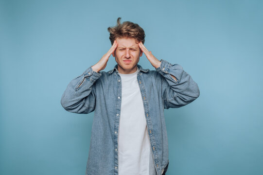 A young man in a denim shirt and white t-shirt stands against a blue background, holding his head with both hands and expressing discomfort or stress.