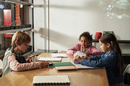 Three children sitting at table working on school assignments, two girls and one boy appearing to be around elementary school age, multiethnic group focused on writing and reading