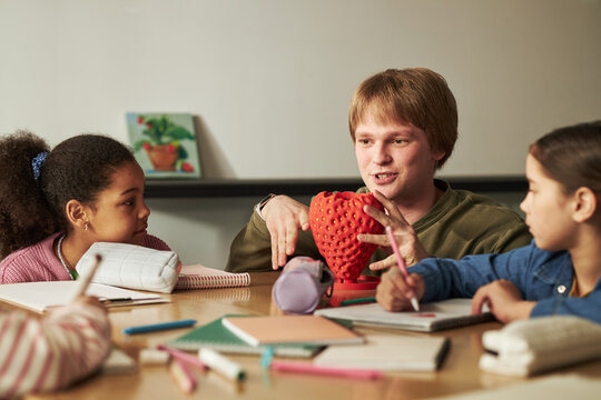 Caucasian young adult man with strawberry model explaining plant growing to diverse group of children sitting around table, children listening attentively and engaging in educational discussion