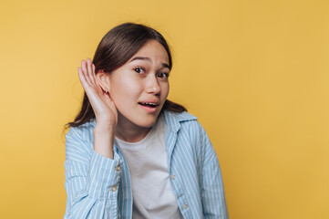 Young woman in a striped shirt holding her hand to her ear, listening intently against a yellow...