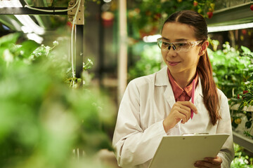 Young Caucasian woman wearing safety glasses working in greenhouse, holding clipboard and...