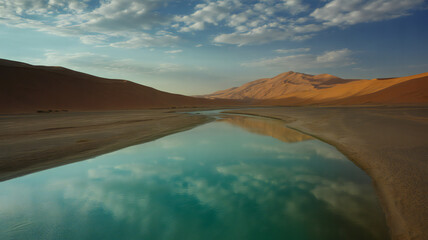 Serene desert oasis reflects dramatic cloudy sky and golden mountain peaks in a vast untouched landscape