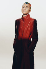 Woman with short colorful hair wearing a long red leather coat, posing confidently against a light...