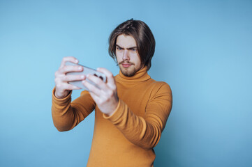 Young man in mustard sweater focused on smartphone against blue background.