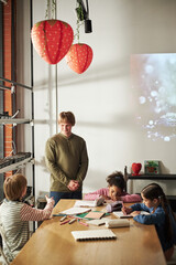 Caucasian male teacher standing near table while diverse group of children sitting and drawing with...