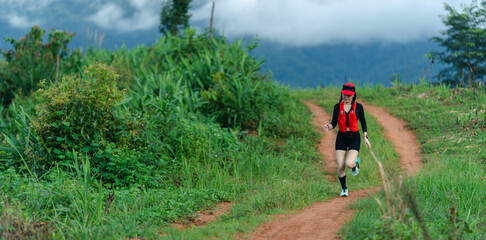 A runner in athletic gear jogging on a dirt trail surrounded by green vegetation under a cloudy sky.