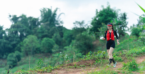 A woman wearing sports gear and a hydration vest walks on a trail through a lush, green forested...